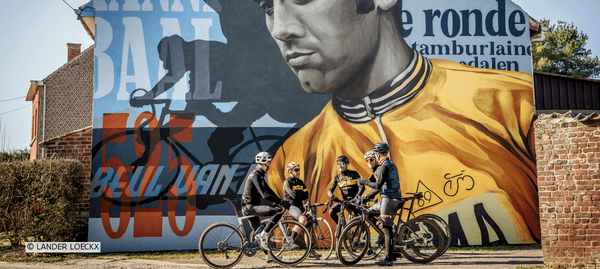 Fietsers staan bij een streetart‑werk ter ere van Eddy Merckx in Kiezegem en praten samen voor het kunstwerk